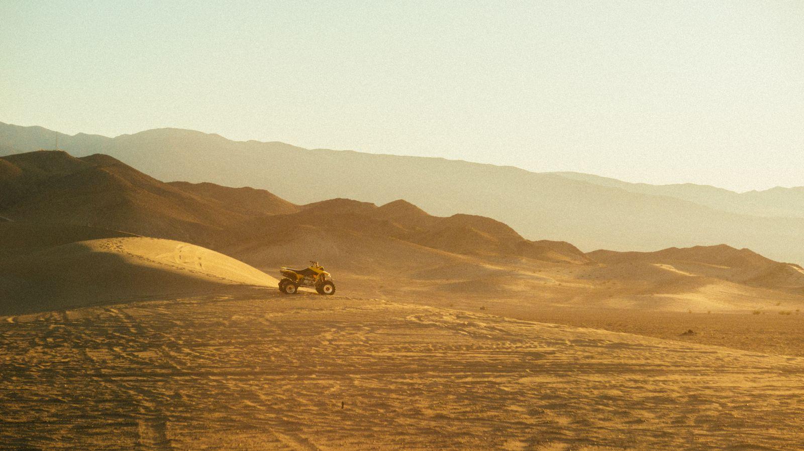 An ATV crossing sunlit desert dunes with mountains in the background