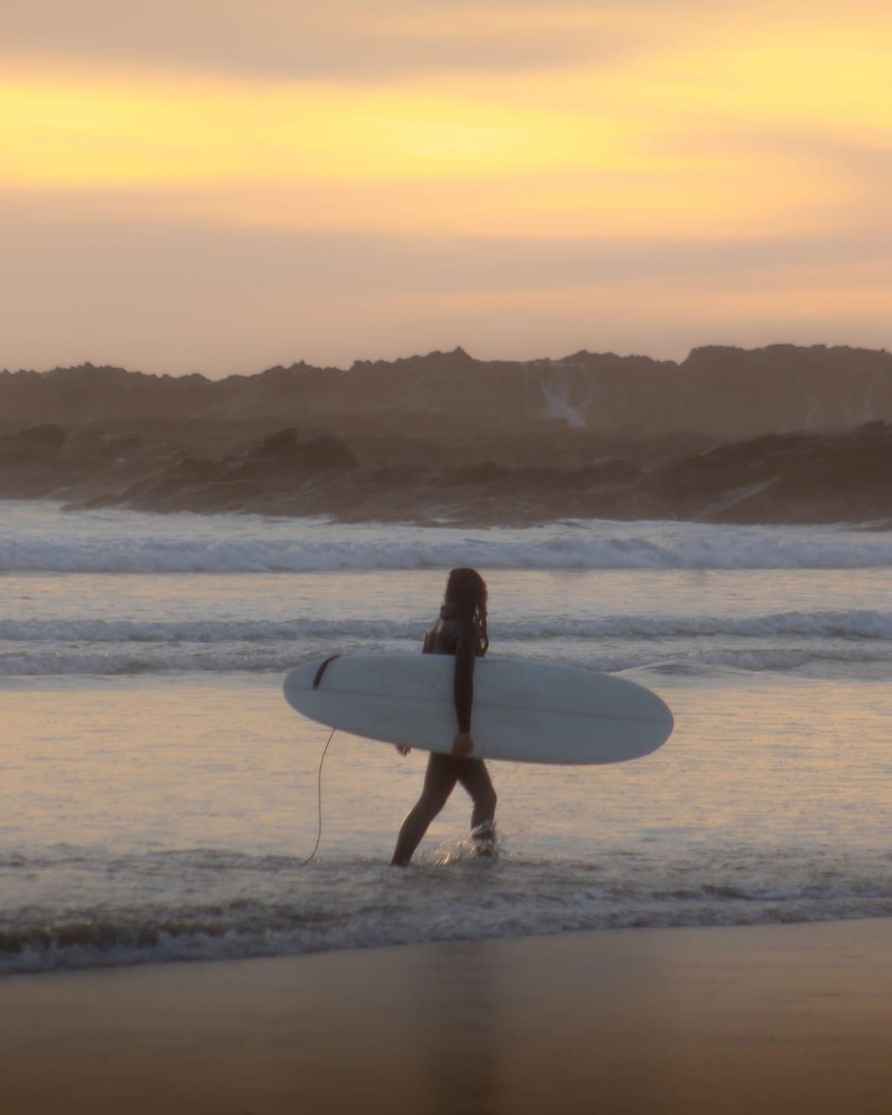 Surfer walking with a board along the shoreline at Playa Los Cerritos at sunset